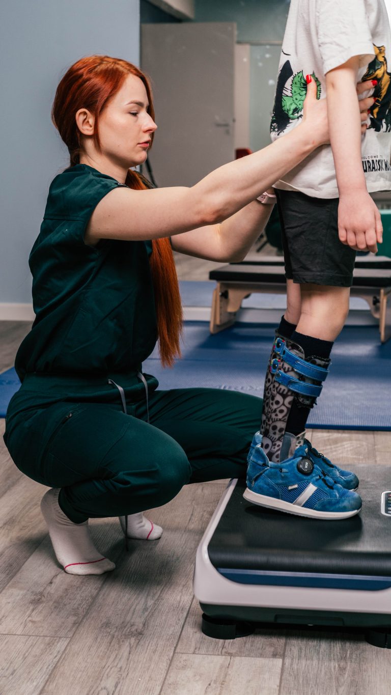 Rehabilitantka w zielonym stroju pomaga dziecku z ortezą na nodze stać na platformie wibracyjnej. Turnus rehabilitacyjny.