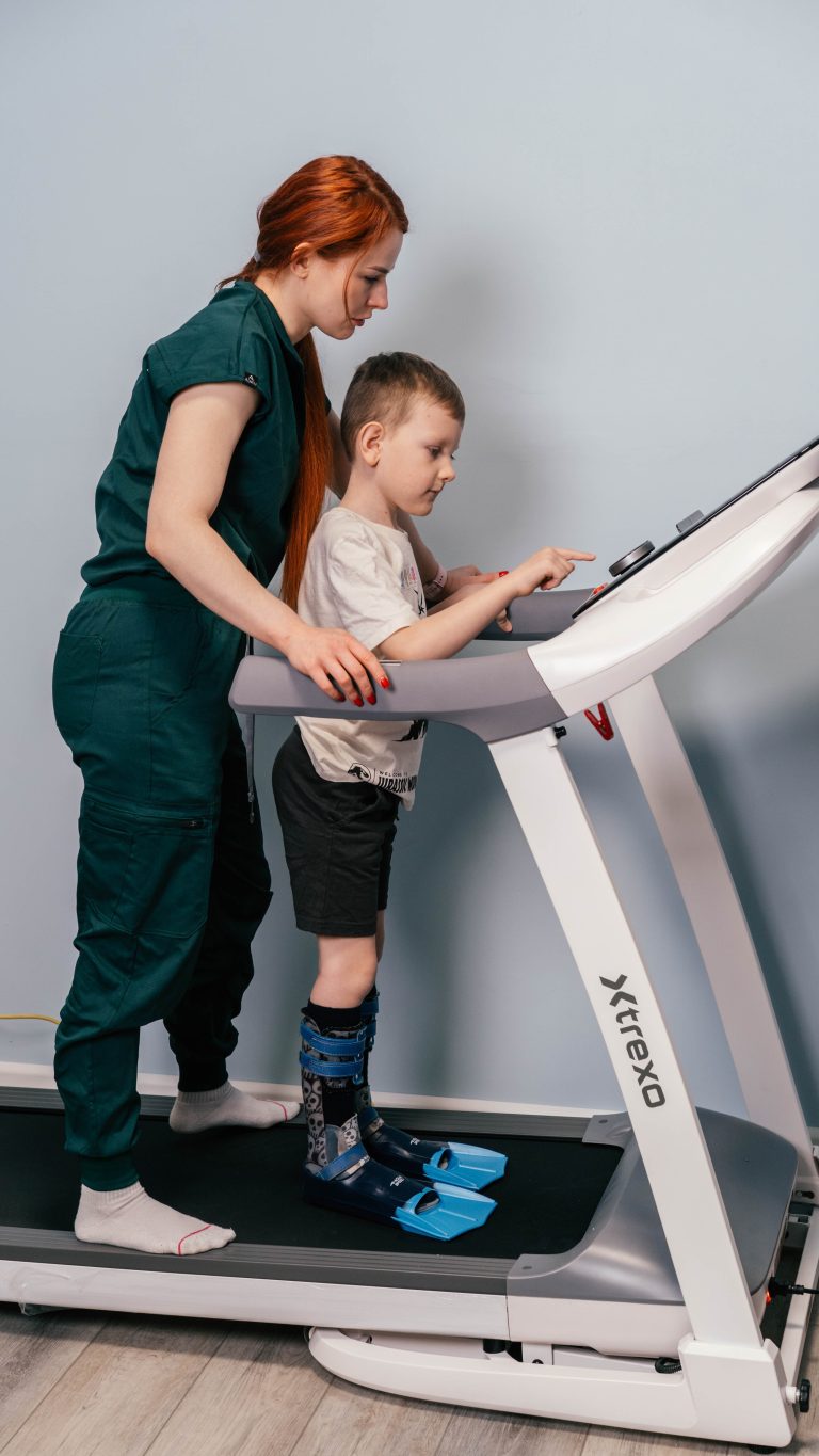 Fizjoterapeutka wspiera dziecko w ortezach na bieżni, które obsługuje panel sterowania podczas turnusu rehabilitacyjnego.
