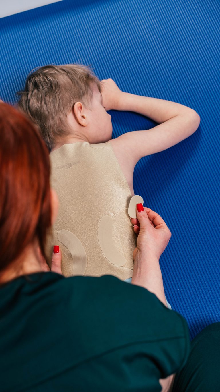 Terapeuta zakłada ortezę tułowia dziecku leżącemu na brzuchu na niebieskiej macie, widoczne plecy dziecka.